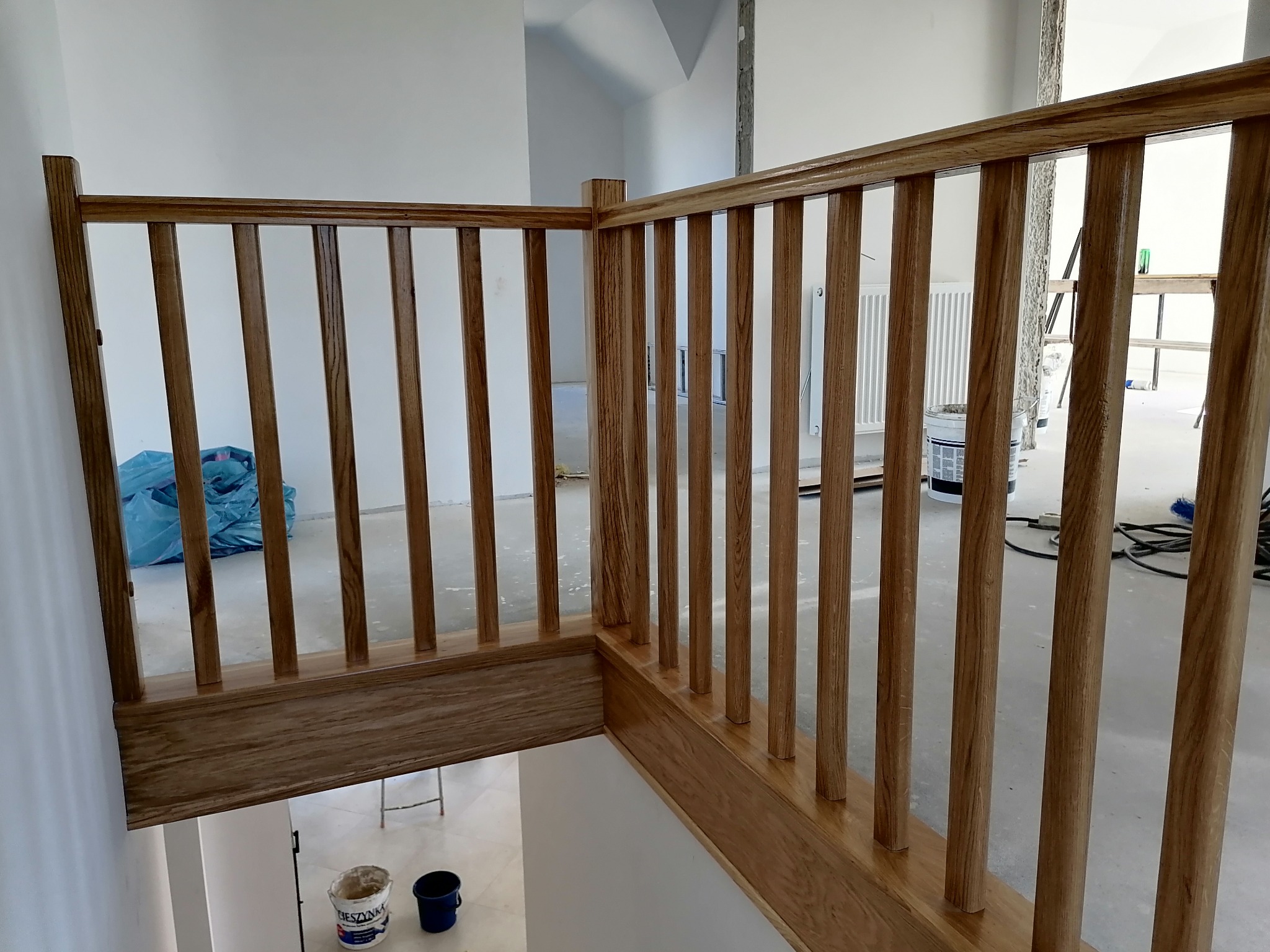 Fragment drewnianej balustrady schodowej z pionowymi szczeblami, widok z góry na białe pomieszczenie w trakcie remontu, z widocznymi wiadrami i narzędziami.