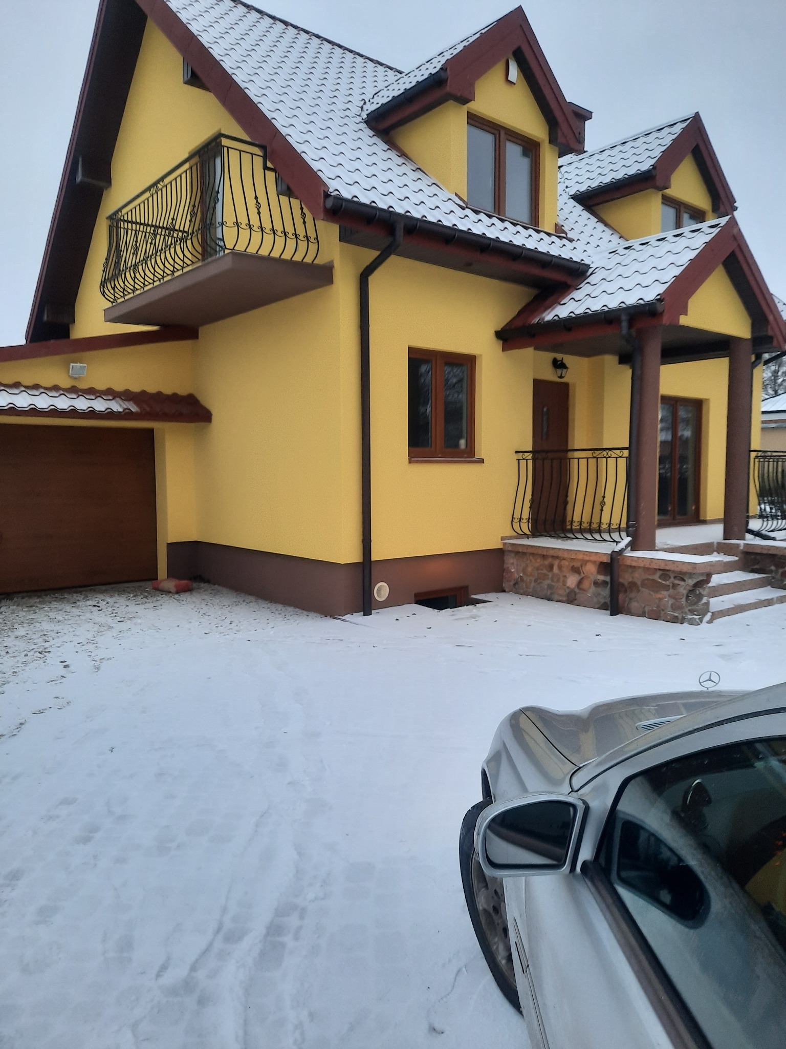 Żółty dom dwukondygnacyjny z brązowymi elementami wykończeniowymi, balkonem z kutą balustradą i śniegiem na dachu, częściowo zasłonięty przez srebrny samochód w zimowej scenerii.