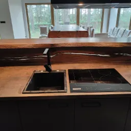 Nowoczesna kuchnia z blatem imitującym rdzę, czarnym zlewem i płytą indukcyjną. Nad blatem drewniana półka z widocznym usłojeniem. W tle stół i krzesła w folii ochronnej oraz widok na las przez...