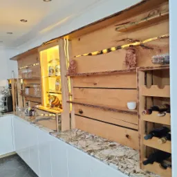 Drewniana zabudowa kuchenna z podświetleniem LED, półkami na szkło i alkohole, oraz rzeźbionymi figurkami, blat z kamienia w kolorze beżowym z białymi żyłkami.