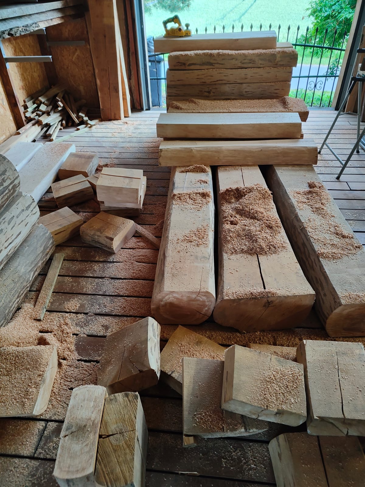 Przygotowane drewniane elementy konstrukcyjne w Wielbrandowie, pokryte wiórami, widoczna strugarka elektryczna w tle.
