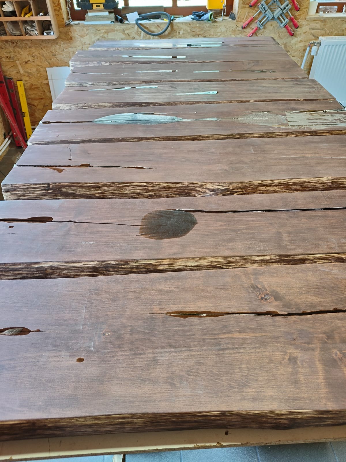 Surowe, brązowe deski drewniane z widocznymi sękami i pęknięciami, ułożone równolegle na stole w warsztacie, czekające na dalszą obróbkę.