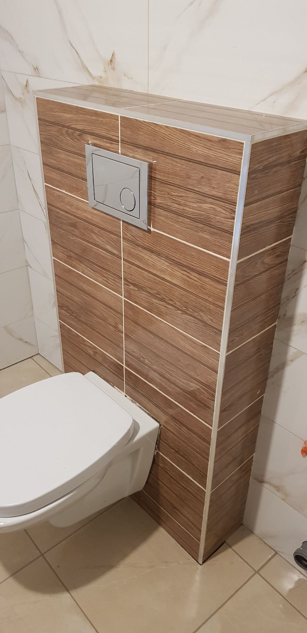 Podwieszana toaleta z zabudową z płytek imitujących drewno, chromowany przycisk spłukiwania, beżowe płytki podłogowe, marmurowe płytki ścienne.