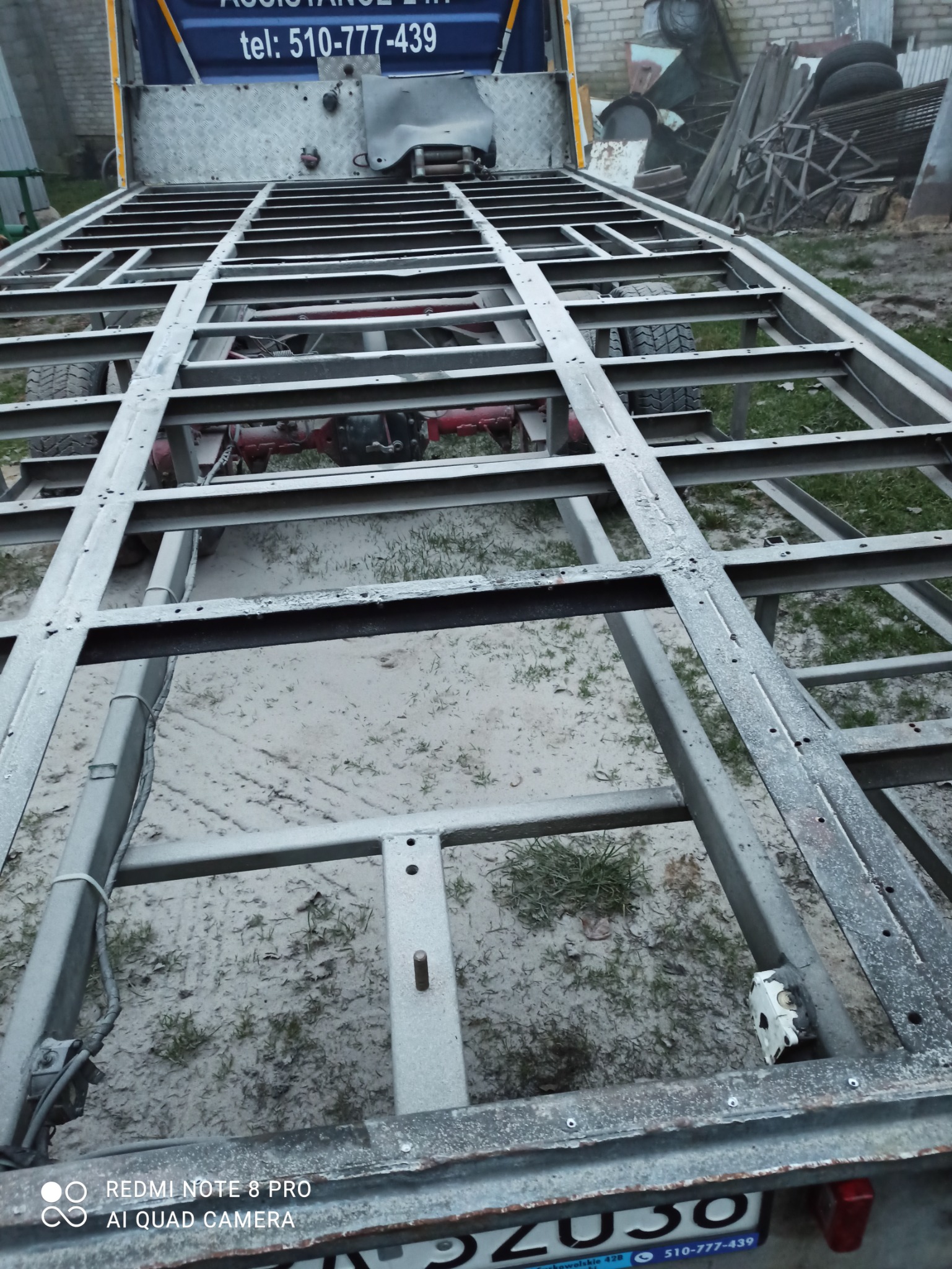 Metalowa rama przyczepy samochodowej po obróbce, widoczne ślady korozji i niedokładnego oczyszczenia, tło z ziemią i trawą.