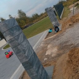 Wyburzenia i Rozbiórki Adam Kuśmierczyk - Betonowy słupek ogrodzeniowy z widoczną strukturą kamienia, osadzony w fundamencie, z zabawką dziecka w tle na świeżo ułożonej kostce brukowej i fragmentem montowanego ogrodzenia panelowego.