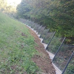 Wyburzenia i Rozbiórki Adam Kuśmierczyk - Ukośne ujęcie metalowego ogrodzenia panelowego na zboczu, z widocznym gruntem i zielenią w tle.