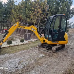Ż&oacute;łta mini koparka JCB 8018 CTS na nier&oacute;wnym, częściowo utwardzonym terenie z widocznymi śladami opon, w tle drzewa iglaste i fragment posesji z elementami drewnianej konstrukcji.