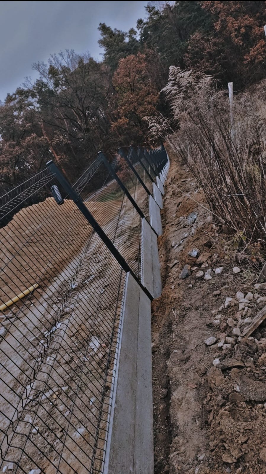 Długi, czarny panelowy system ogrodzeniowy z betonową podmurówką, zainstalowany na nierównym, zalesionym terenie ze stromym zboczem.
