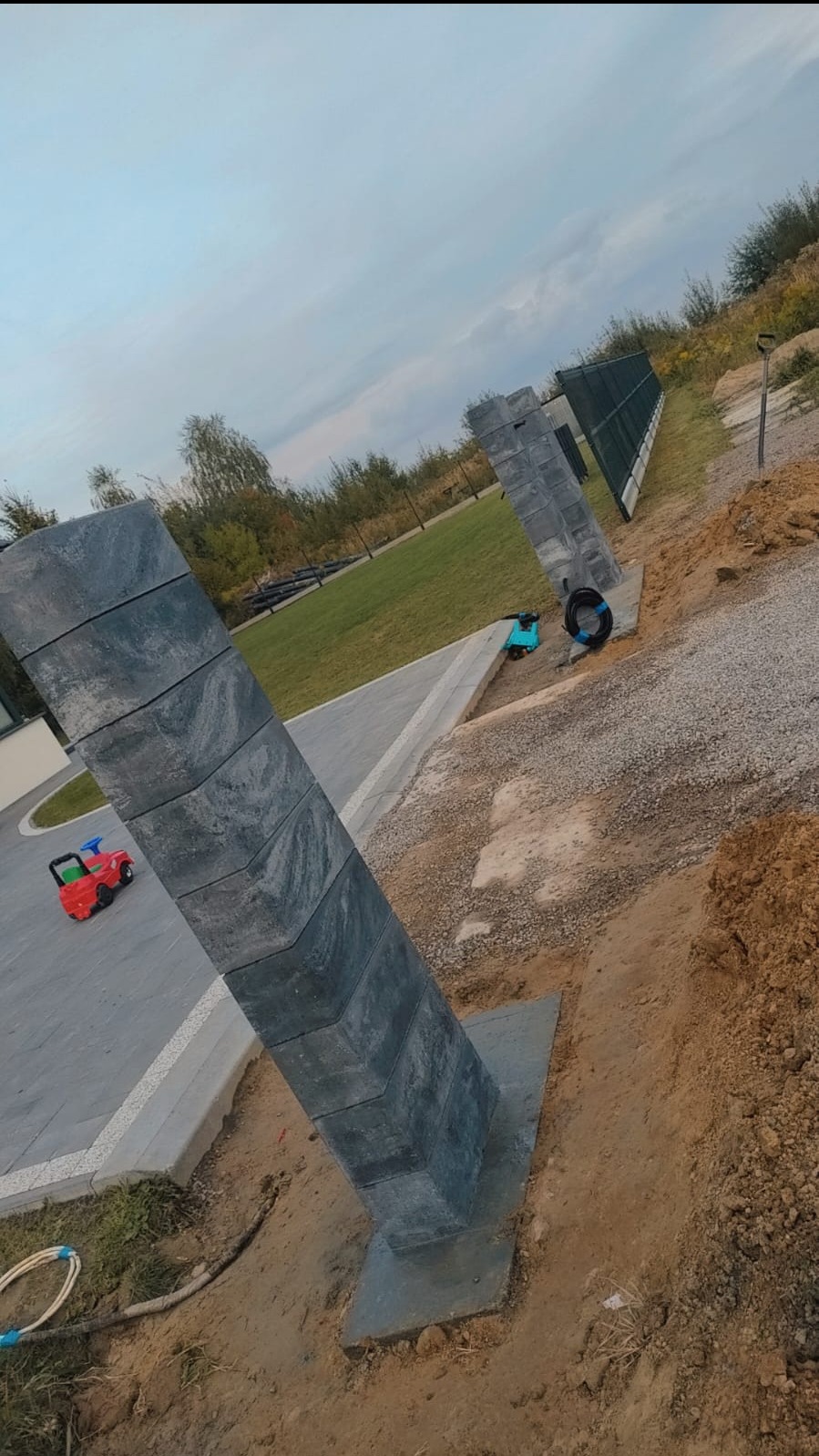 Betonowy słupek ogrodzeniowy z widoczną strukturą kamienia, osadzony w fundamencie, z zabawką dziecka w tle na świeżo ułożonej kostce brukowej i fragmentem montowanego ogrodzenia panelowego.