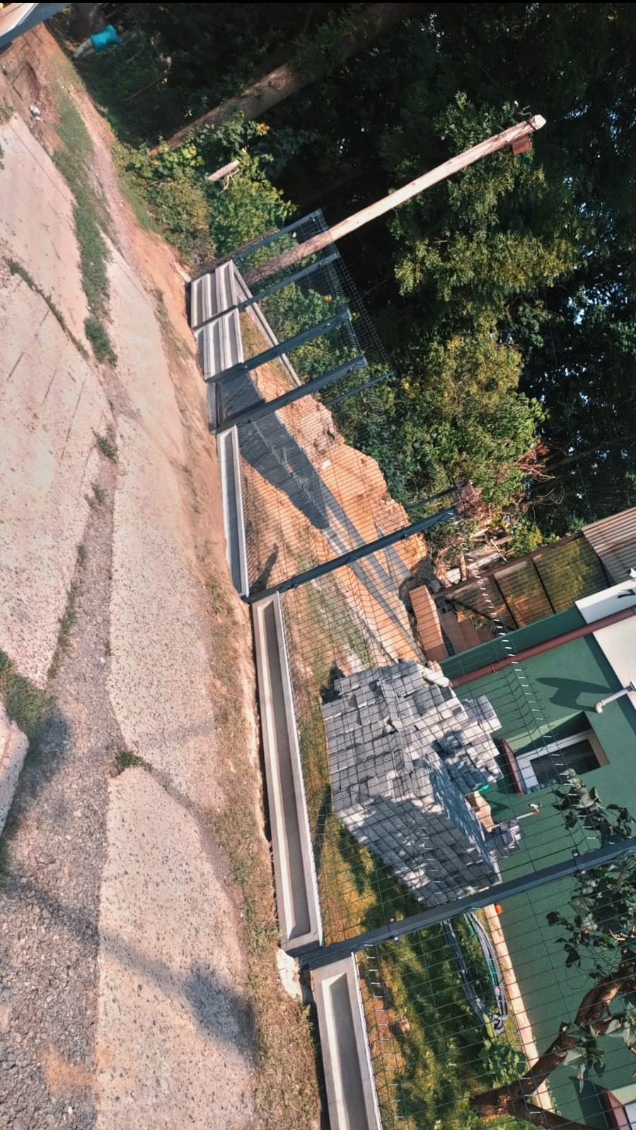 Montaż ogrodzenia panelowego z podmurówką prefabrykowaną, widoczne stosy kostki brukowej przygotowane do układania.