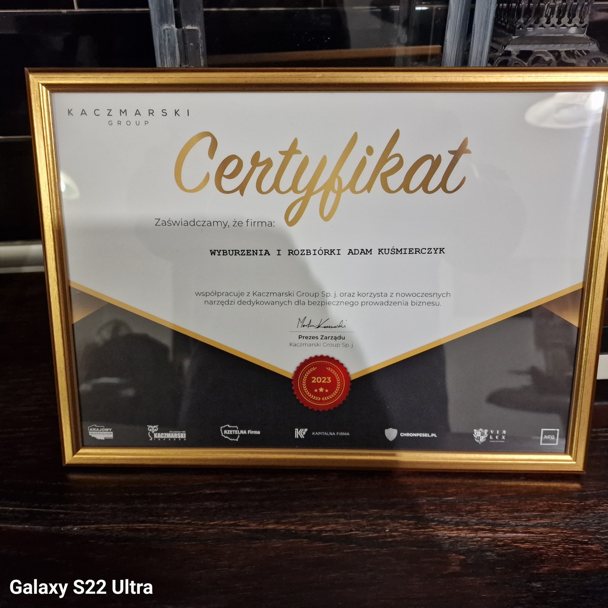 Oprawiony w złotą ramkę certyfikat firmy Kaczmarski Group, potwierdzający współpracę i korzystanie z nowoczesnych narzędzi w obszarze wyburzeń i rozbiórek, sygnowany przez Prezesa Zarządu...