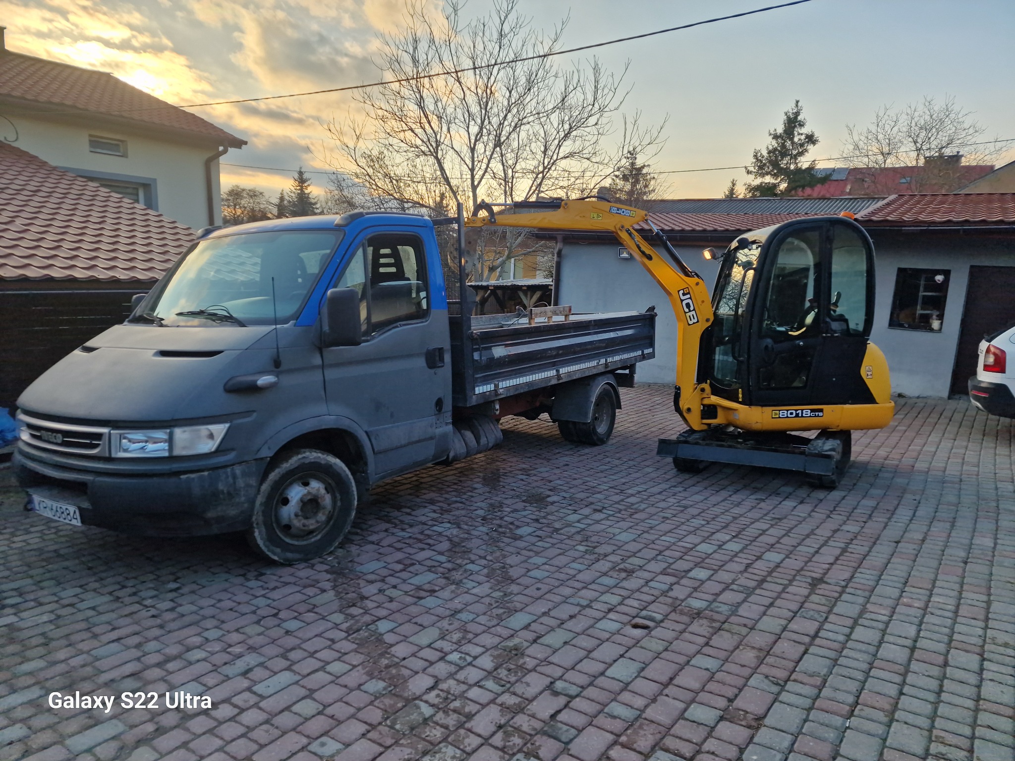 Szaro-niebieska ciężarówka Iveco z otwartą skrzynią ładunkową i żółta minikoparka JCB 8018CTS na kostce brukowej przed budynkiem w słoneczny dzień.