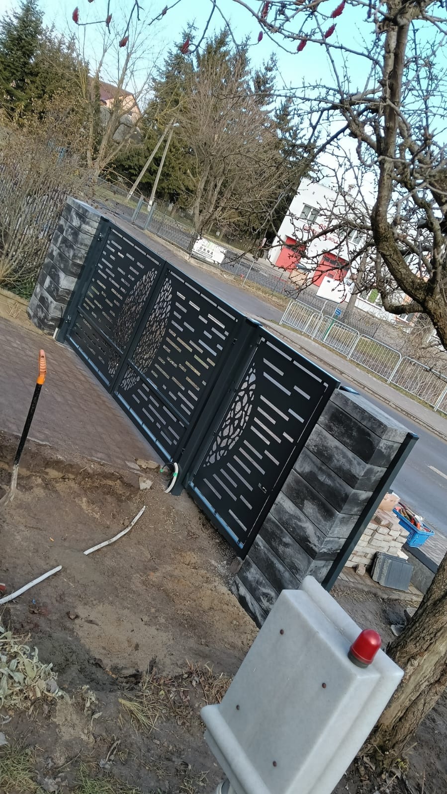 Nowoczesna brama wjazdowa z metalowymi przęsłami o ażurowym wzorze, osadzona na słupkach z szarej kostki, z widocznym automatem do bramy i elementami przygotowanymi do montażu, na tle ulicy...