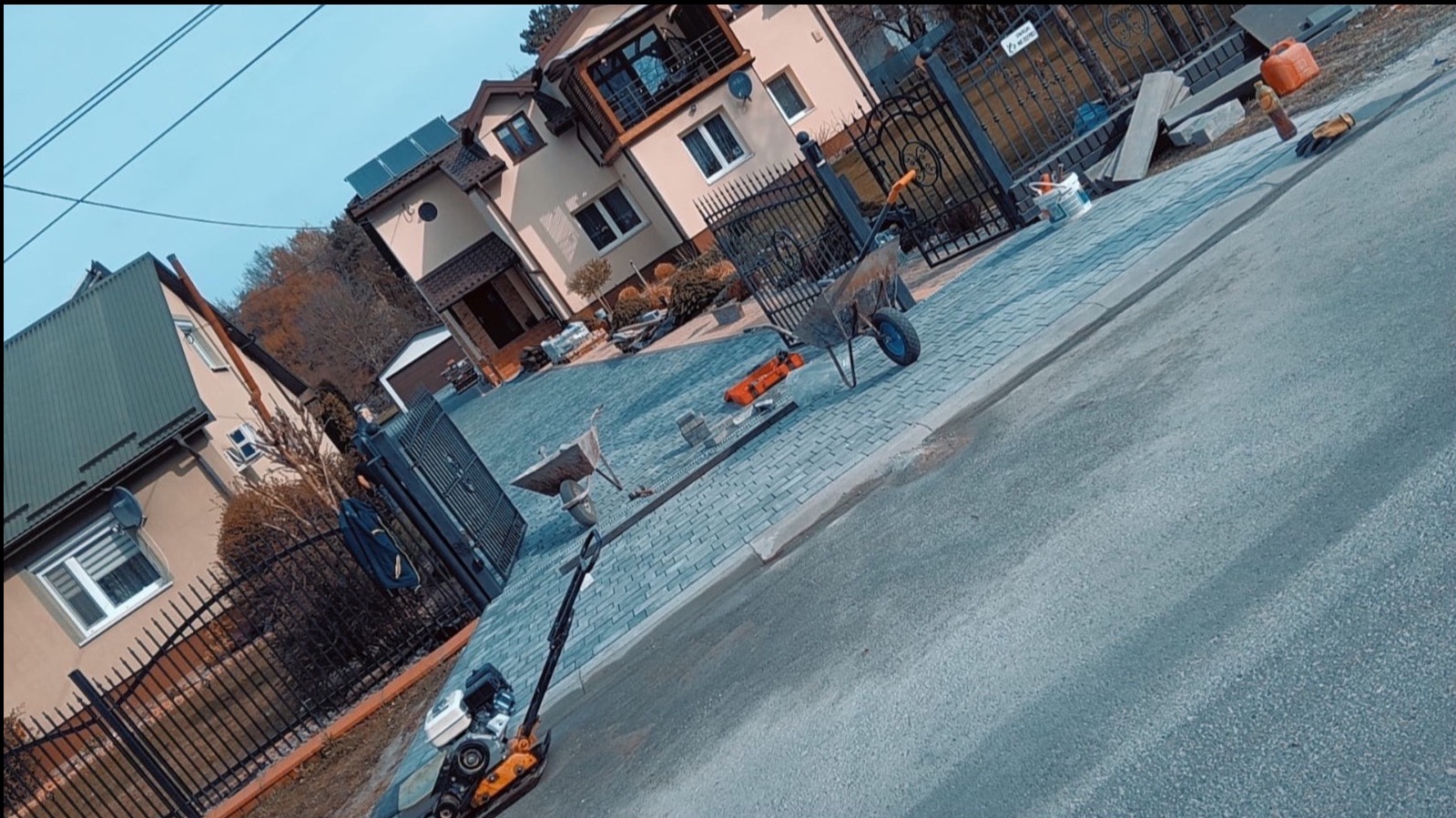 Ułożona kostka brukowa na podjeździe domu jednorodzinnego, widoczne narzędzia i maszyny budowlane, metalowe ogrodzenie, brama. Kąt ujęcia z dołu.