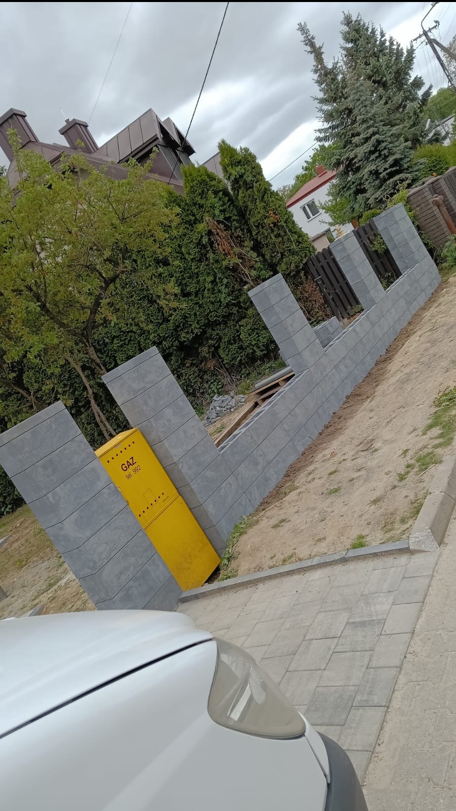 Szara, modułowa konstrukcja ogrodzenia w trakcie budowy, z widoczną żółtą skrzynką gazową. Częściowo ułożona kostka brukowa i fragment białego samochodu w kadrze.