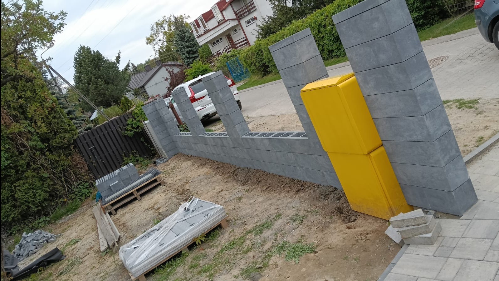 Wznoszenie ogrodzenia z szarej, betonowej kostki modułowej. Widoczna żółta skrzynka pocztowa wkomponowana w konstrukcję. Częściowo ułożona kostka brukowa na podjeździe.