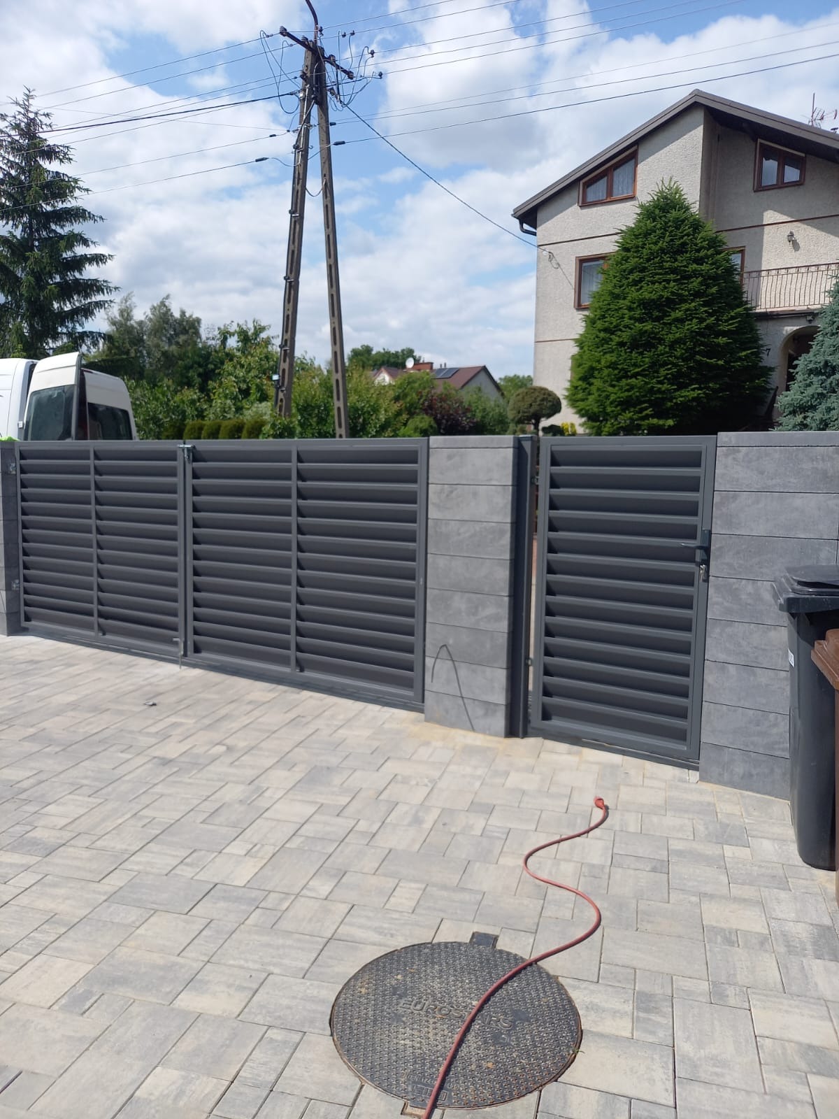 Nowoczesna, szara brama wjazdowa z poziomymi lamelami, osadzona na murowanych słupkach z szarej kostki. Widok na posesję z kostką brukową i studzienką.