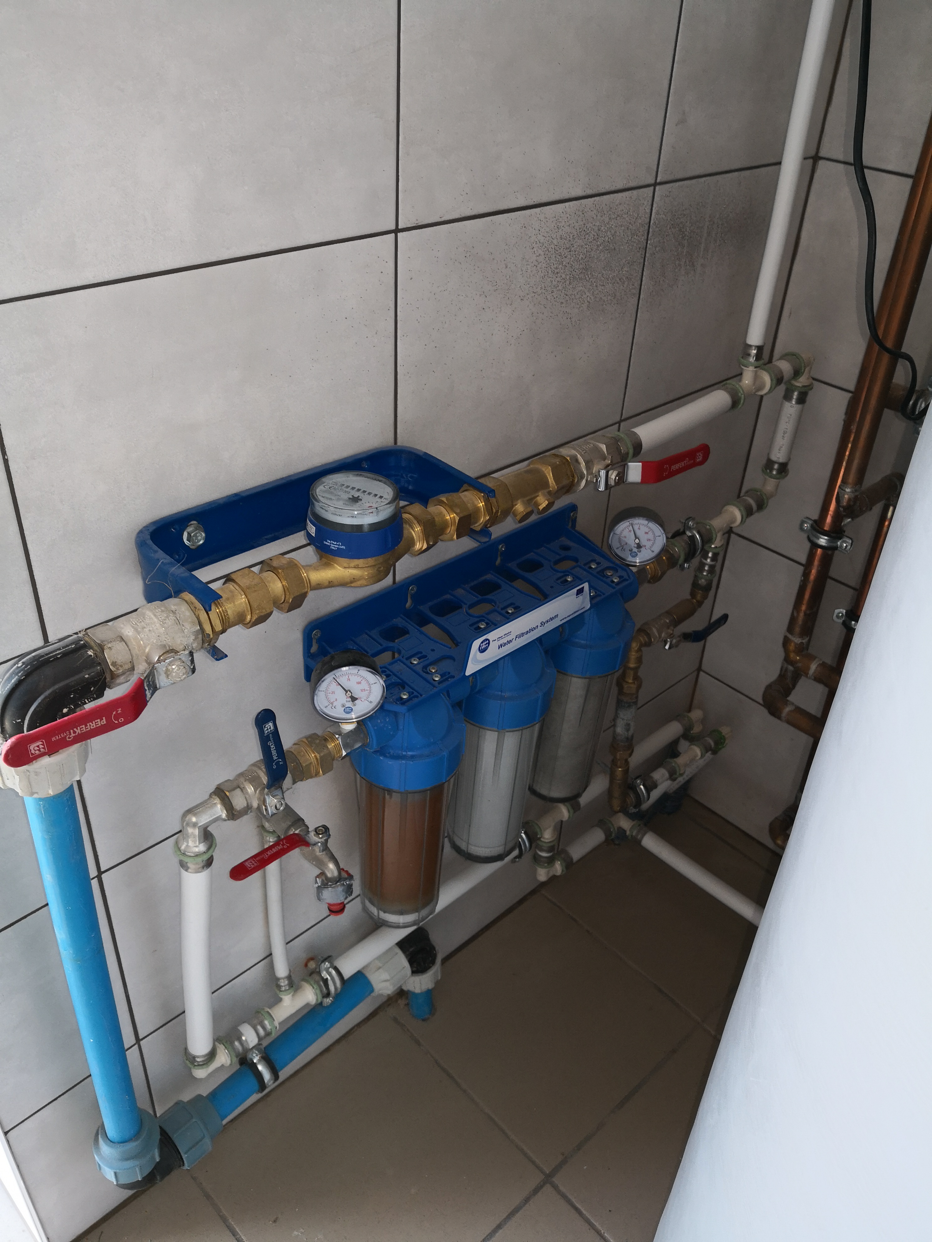 Zestaw filtrów wody zamontowany na ścianie z szarych płytek, widoczne manometry, zawory kulowe z czerwonymi rączkami i rury z tworzyw sztucznych.