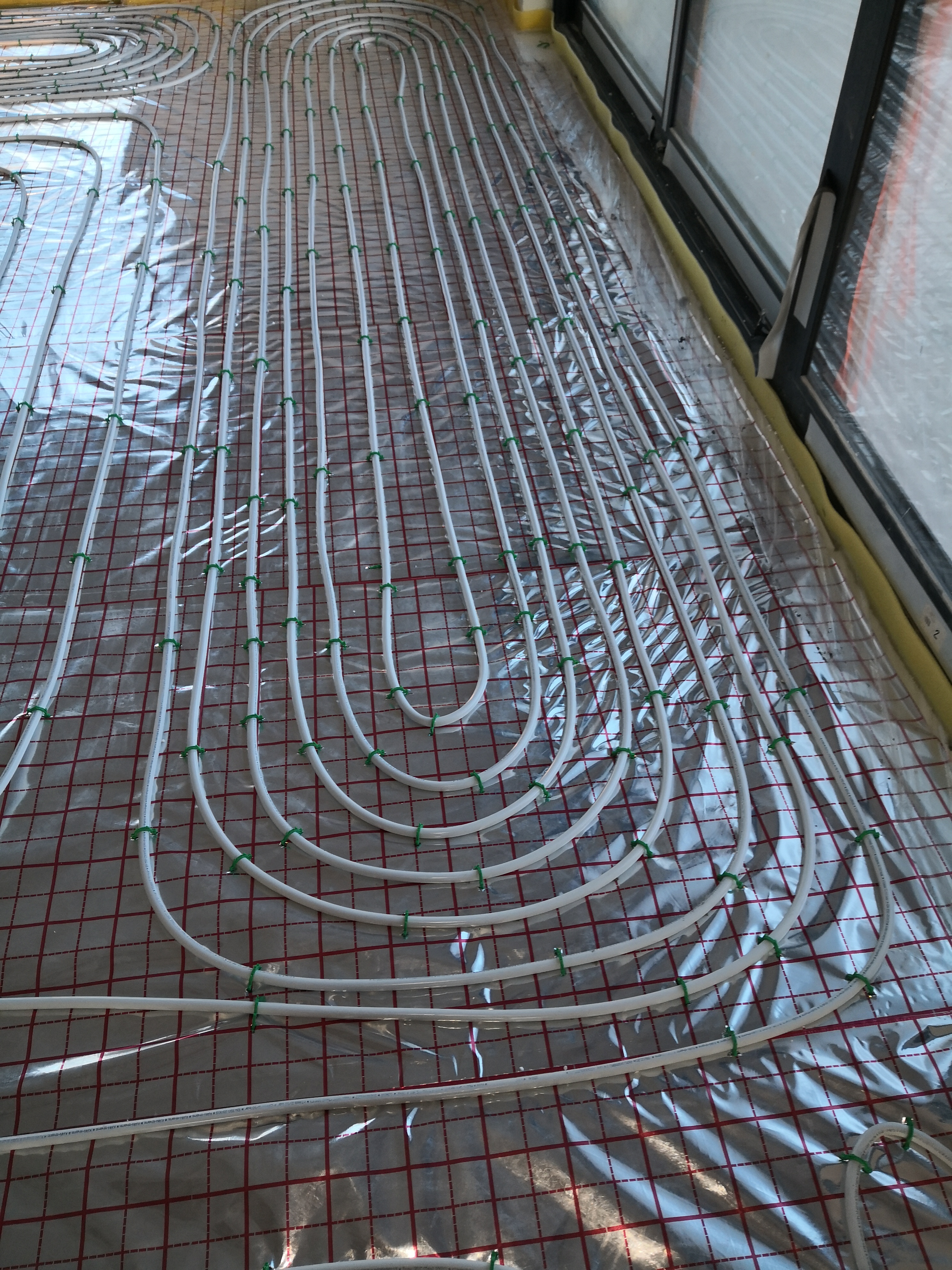 Białe rury ogrzewania podłogowego ułożone w pętle na folii z czerwoną siatką, przymocowane zielonymi klipsami, widoczne okno w tle.