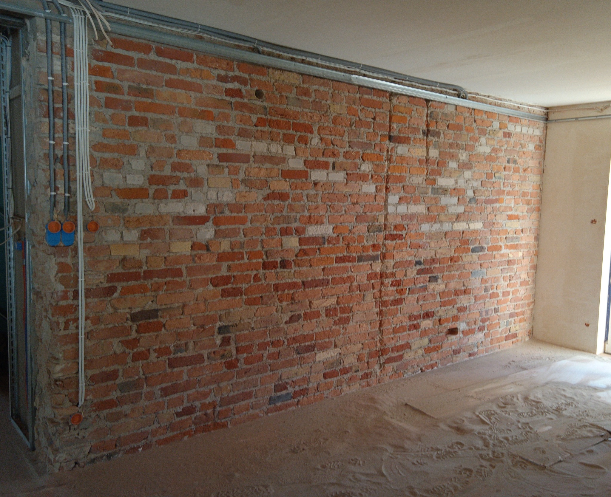 Surowa ściana z czerwonej cegły z widocznymi instalacjami elektrycznymi w trakcie remontu, z metalowymi profilami pod sufitem i piaskiem na podłodze.