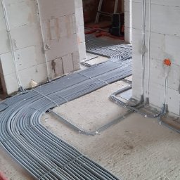 INEL-TEL Grzegorz Libera - Szare rury instalacji elektrycznej ułożone na podłodze w surowym wnętrzu budynku w trakcie budowy. Widoczne puszki elektryczne w ścianach i drabina w tle.