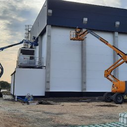 INEL-TEL Grzegorz Libera - Dwie podnośniki koszowe przy montażu oświetlenia zewnętrznego na dużej hali. Jeden pomarańczowy, drugi niebieski. Widoczne lampy na elewacji.