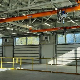 INEL-TEL Grzegorz Libera - Wnętrze hali produkcyjnej z suwnicami pod stropem i oknami. Widoczne bramy segmentowe, oświetlenie przemysłowe i żółte barierki ochronne.