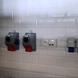 INEL-TEL Grzegorz Libera - Rząd gniazd przemysłowych i standardowych na białej, kafelkowej ścianie. Widoczne różne typy gniazd, w tym gniazda siłowe z czerwonymi i niebieskimi elementami.