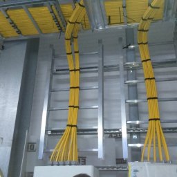 INEL-TEL Grzegorz Libera - Żółte kable elektryczne ułożone w korytach kablowych, przymocowane czarnymi opaskami, widoczne elementy konstrukcji metalowej i fragment kasku ochronnego.