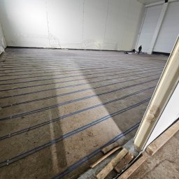 INEL-TEL Grzegorz Libera - Układanie przewodów grzewczych w hali w Rzeszowie. Widoczne równoległe niebieskie rurki na betonowej posadzce, przygotowane do zalania. Prace instalacyjne w toku.