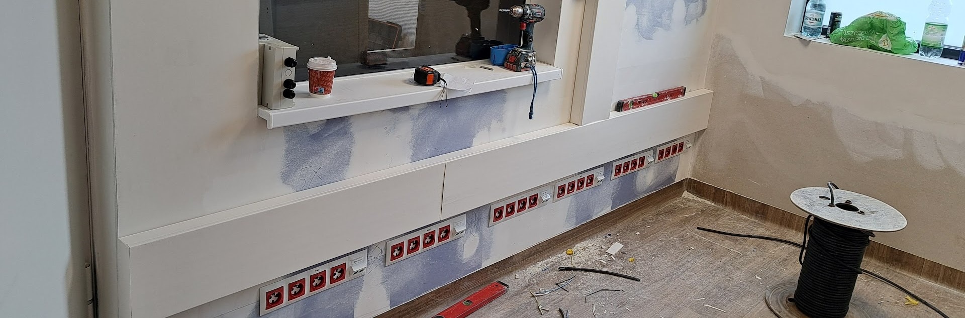 Rząd czerwonych gniazdek z włącznikami zamontowany na ścianie, widoczne narzędzia pracy, poziomica, przewody, wiertarka. Ściana przygotowana do malowania.