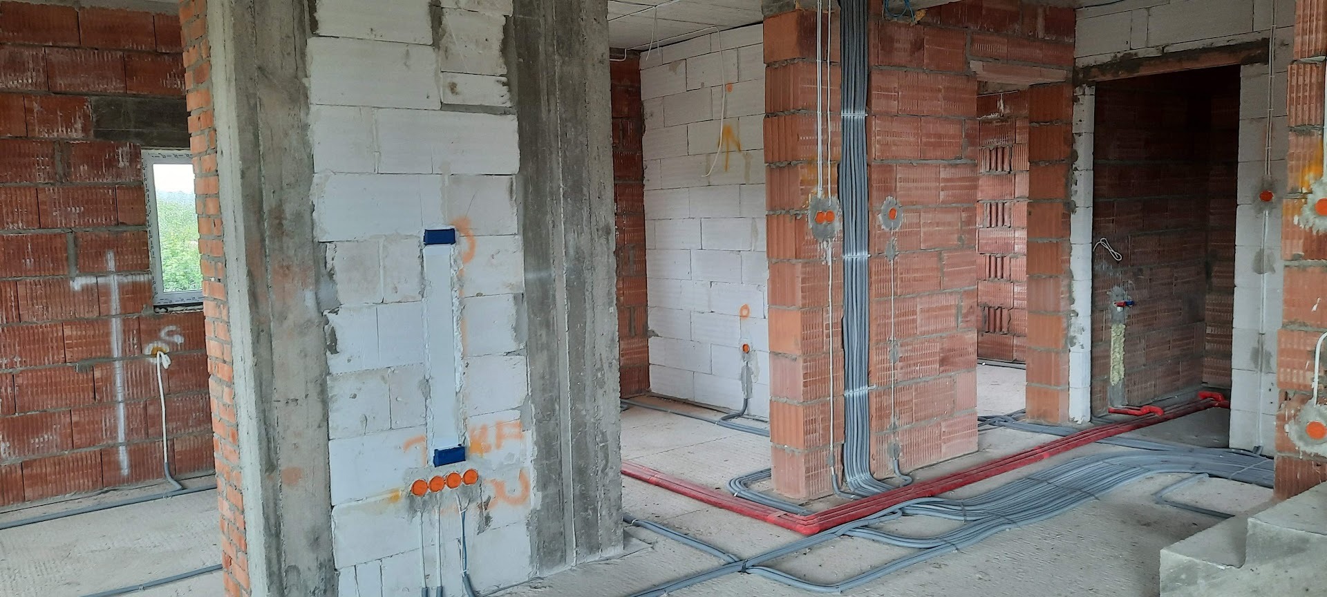 Wnętrze w trakcie budowy z widoczną instalacją elektryczną: szare kable prowadzone wzdłuż ścian z czerwonej cegły i białego bloczka, puszki elektryczne w ścianach.