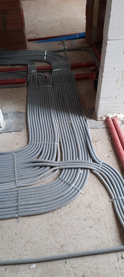 Szare rury instalacyjne ułożone równolegle na podłodze w surowym stanie, zabezpieczone opaskami, z widocznymi fragmentami ścian z cegły i bloczków.