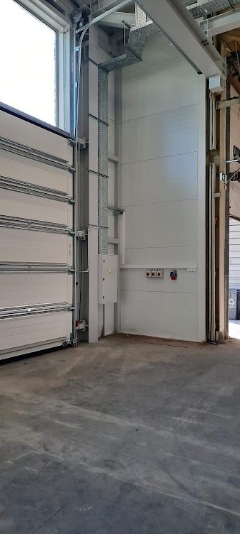 Biała skrzynka elektryczna z przewodami i metalowymi korytkami przymocowana do ściany w hali z otwartą bramą segmentową. Widoczne gniazda i włączniki.