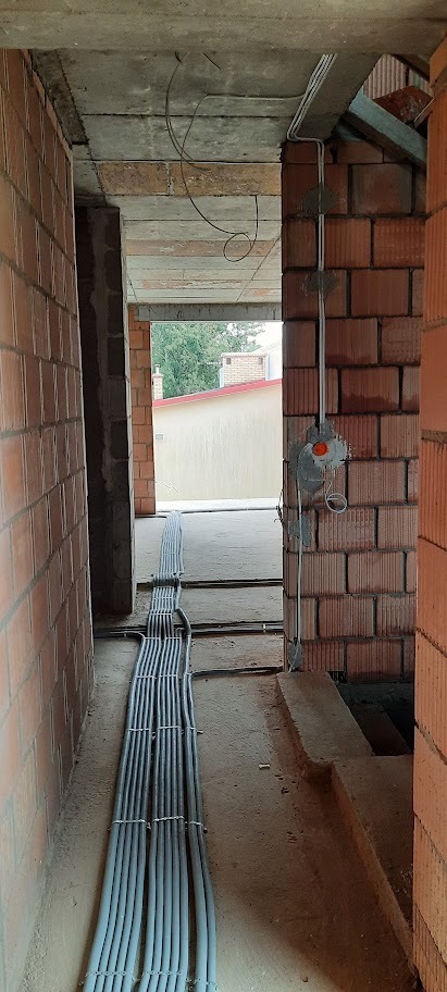 Szereg szarych rur instalacyjnych ułożonych równolegle na podłodze w surowym wnętrzu budynku z czerwonej cegły. Widoczne puszki instalacyjne na ścianie.