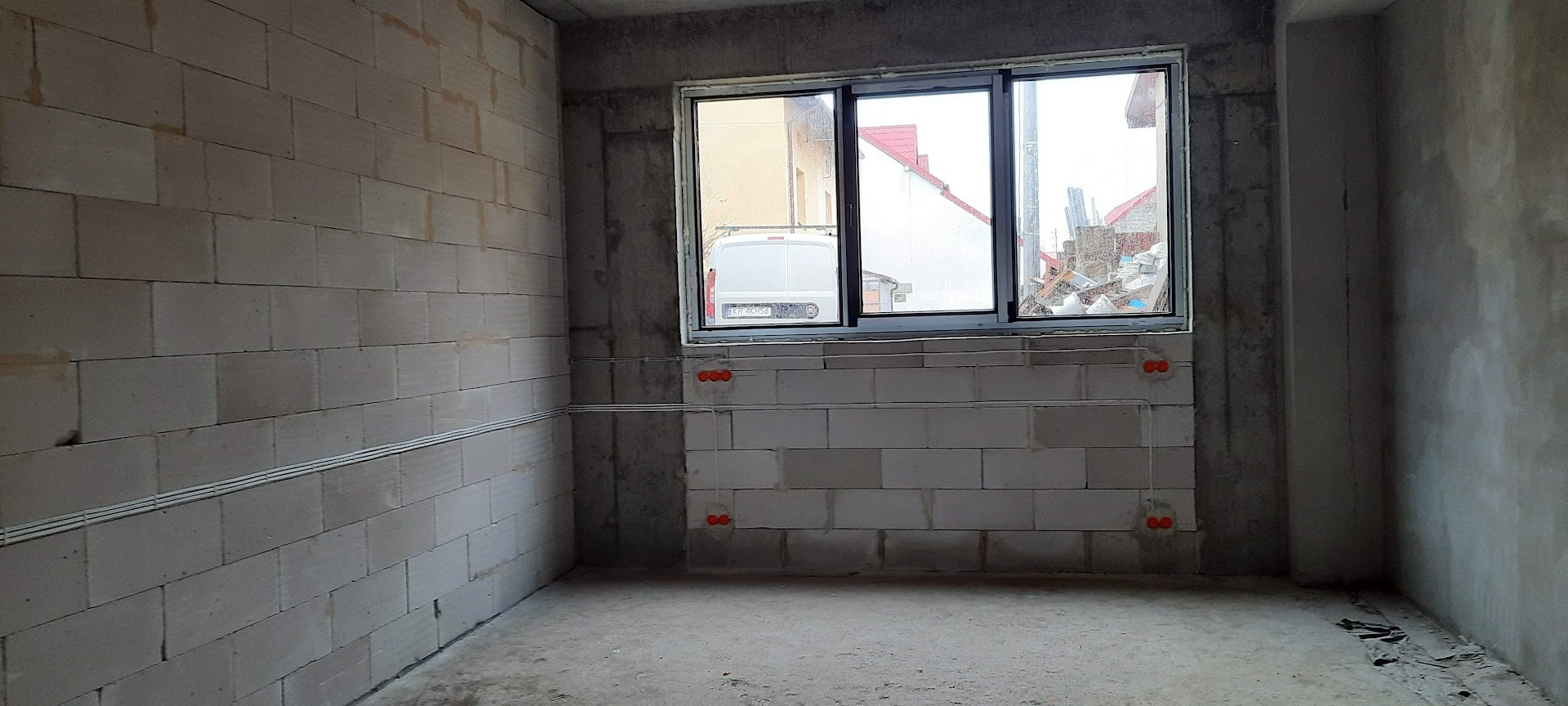 Wnętrze w stanie surowym z widoczną instalacją elektryczną w peszlach i puszkami podtynkowymi. Okno z widokiem na podwórze z zaparkowanym busem.