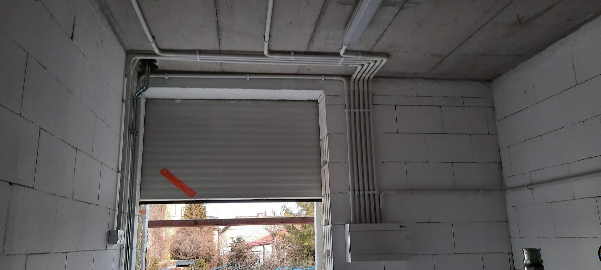 Surowy stan: białe rury instalacji elektrycznej prowadzone po ścianach z betonu komórkowego, widoczna skrzynka rozdzielcza i brama garażowa z oknem na zewnątrz.