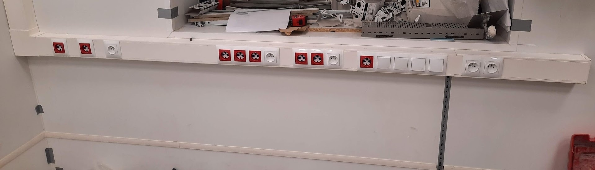 Biała listwa przypodłogowa z zamontowanymi gniazdkami elektrycznymi (czerwone i białe) oraz włącznikami światła na białej ścianie. Nad listwą widoczne materiały budowlane.