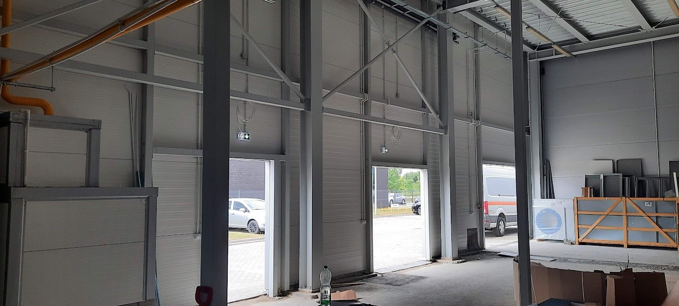 Wnętrze hali przemysłowej z widocznymi instalacjami pod sufitem, stalową konstrukcją i oświetleniem awaryjnym nad otwartymi drzwiami. Na zewnątrz widoczne pojazdy.