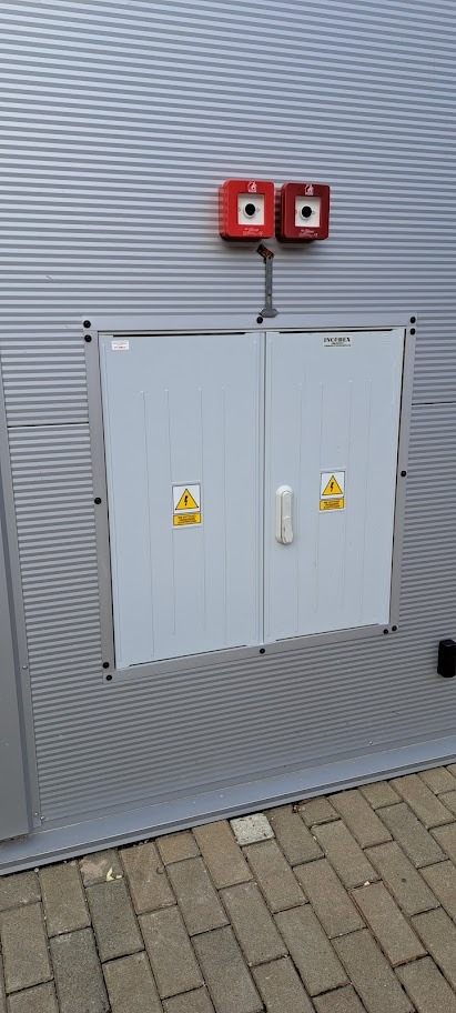 Szara szafa elektryczna z dwoma czerwonymi sygnalizatorami po bokach, umieszczona na metalowej ścianie z poziomymi żłobieniami. Na drzwiach szafy żółte znaki ostrzegawcze.