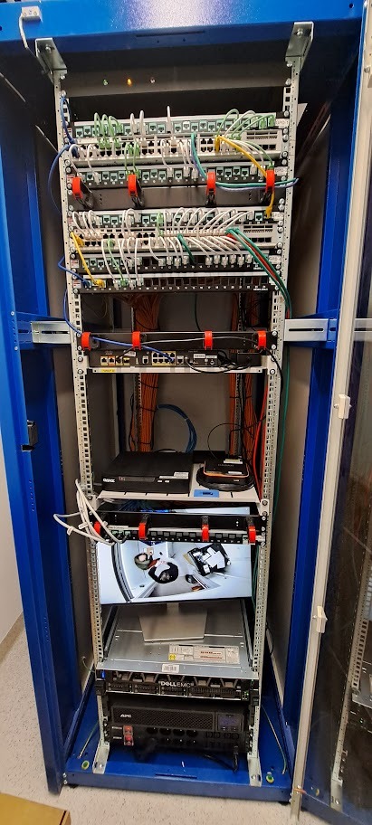 Niebieska szafa rack z zamontowanymi urządzeniami sieciowymi, patch panele, switche, serwery. Widoczne kable i organizatory kabli. Dolny ekran wyświetla obraz z kamery.