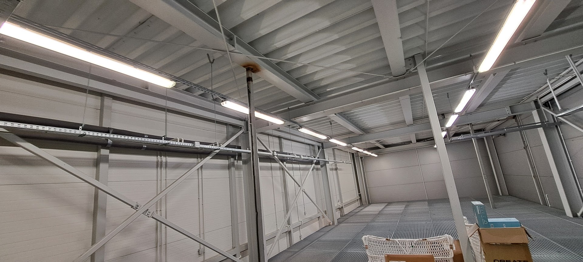 Wnętrze hali przemysłowej z zamontowanym oświetleniem liniowym. Widoczne metalowe konstrukcje wsporcze i instalacje pod sufitem. Oświetlenie równomiernie rozmieszczone.