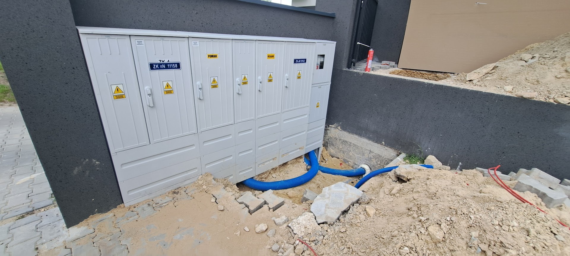 Szara szafa elektryczna z niebieskimi rurami osłonowymi na zewnątrz budynku. Widoczne oznaczenia ostrzegawcze i numery identyfikacyjne na drzwiach szafy.