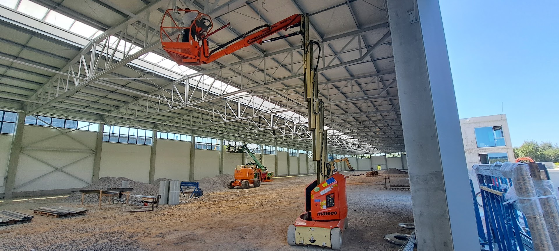 Wnętrze hali w trakcie budowy z podnośnikami koszowymi i pracownikami na wysokości, widoczne elementy konstrukcyjne dachu i ścian. Jasne oświetlenie dzienne.