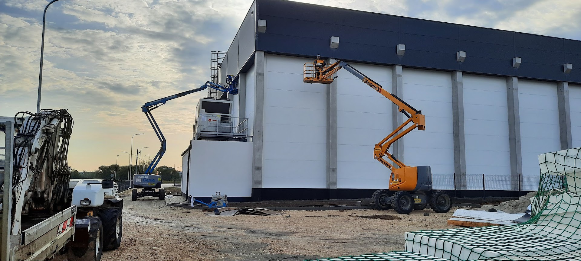 Dwie podnośniki koszowe na budowie hali. Jeden pomarańczowy z pracownikiem, drugi niebieski. W tle błękitne niebo z chmurami.
