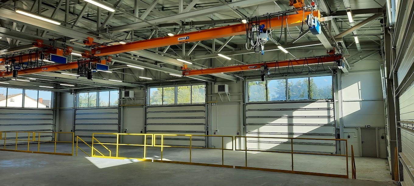 Wnętrze hali produkcyjnej z suwnicą ABUS, widoczne okna, bramy segmentowe i żółte barierki. Oświetlenie sufitowe. Hala w jasnych barwach.