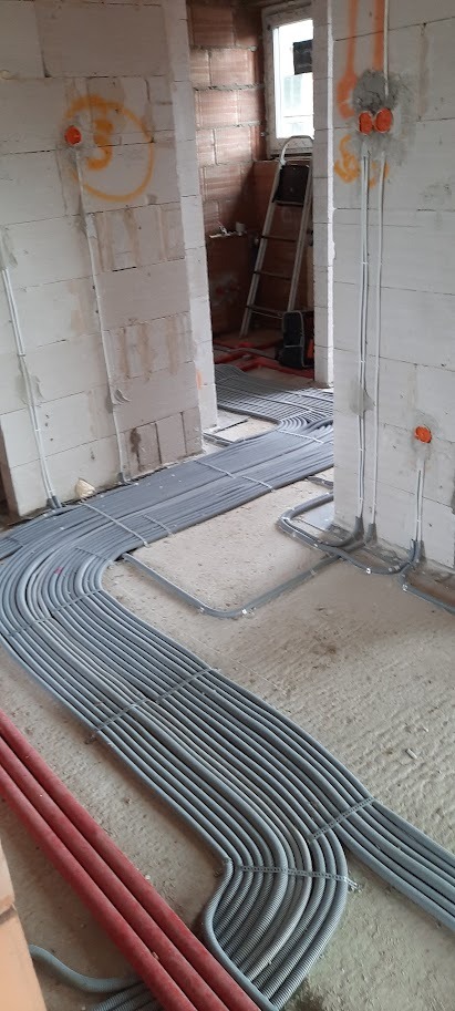 Szare rury instalacji elektrycznej ułożone na podłodze w surowym wnętrzu budynku w trakcie budowy. Widoczne puszki elektryczne w ścianach i drabina w tle.