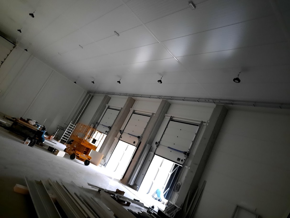 Wnętrze hali magazynowej z zamontowanym oświetleniem sufitowym. Widoczny podnośnik koszowy i częściowo otwarte bramy segmentowe. Surowy stan wykończenia.