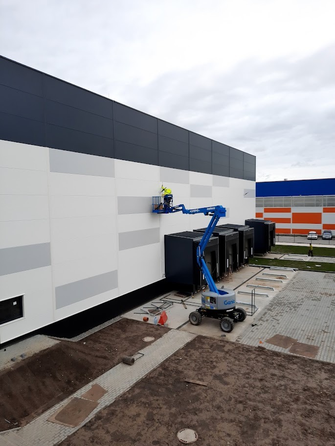 Pracownik w podnośniku koszowym montuje instalację na elewacji hali magazynowej. Widoczne elementy instalacji, narzędzia i fragment terenu wokół budynku.