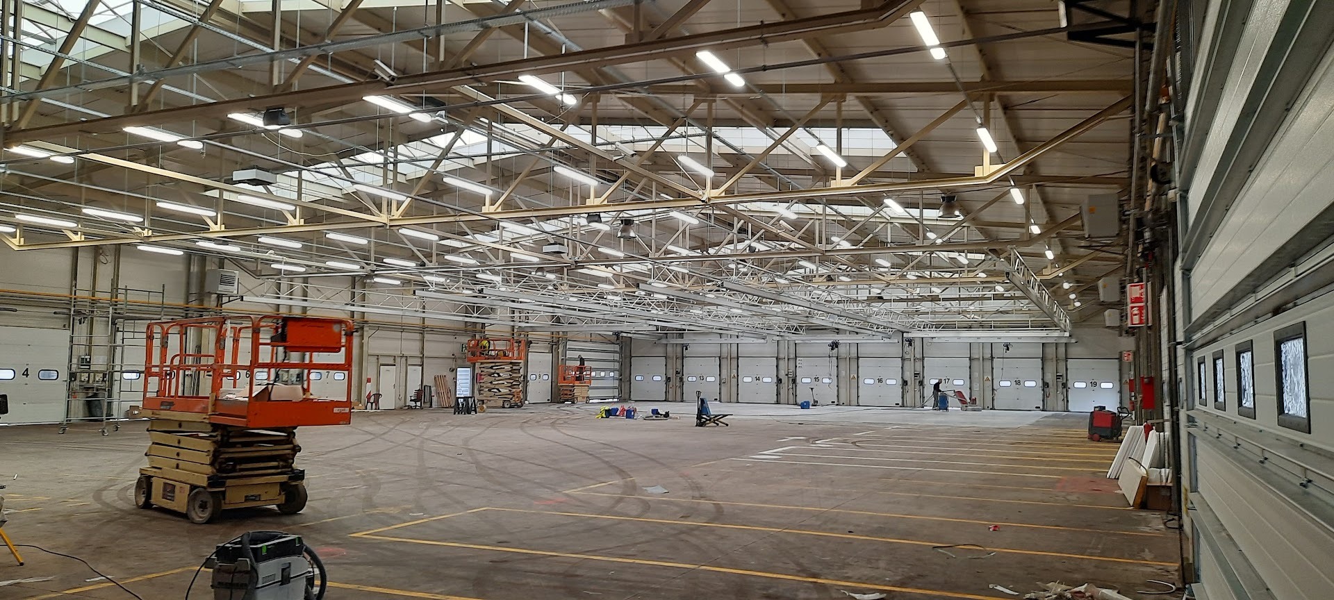 Montaż oświetlenia w hali magazynowej z użyciem podnośników. Widoczne lampy, konstrukcja dachu, brama garażowa i sprzęt instalacyjny.